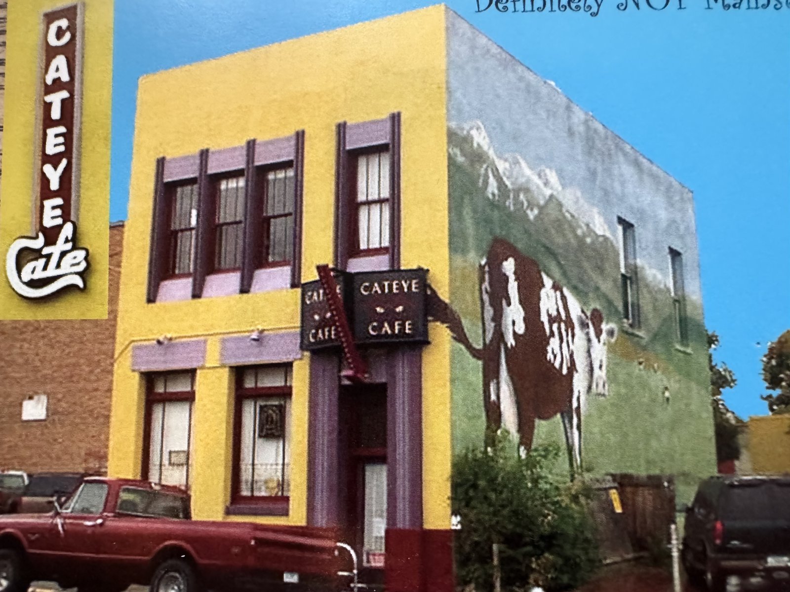 Cat Eye Café’s yellow building and neon sign beside a painted cow mural