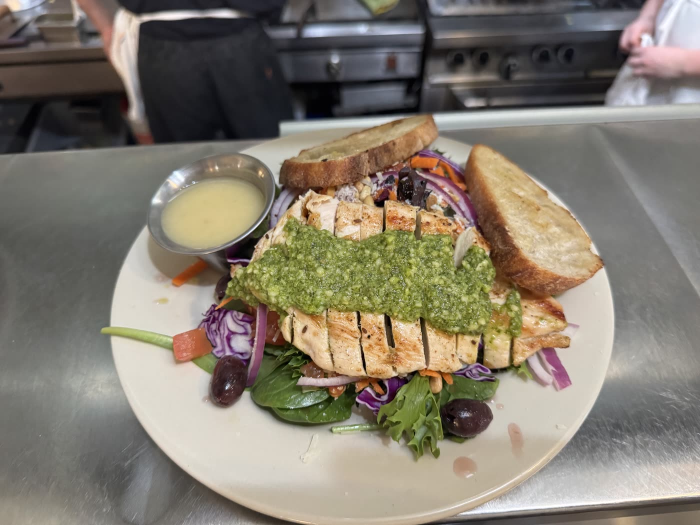 Sliced grilled chicken with pesto over greens, olives and purple cabbage, with toasted bread and dressing